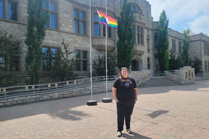 Image of Jocelyn Ormerod standing on USask campus in from of Pride flag
