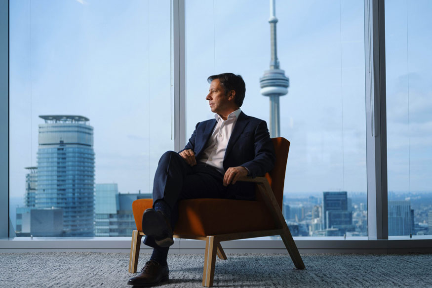 Kevin Peesker sitting on chair in front of floor to ceiling high-rise window over-looking Toronto skyline