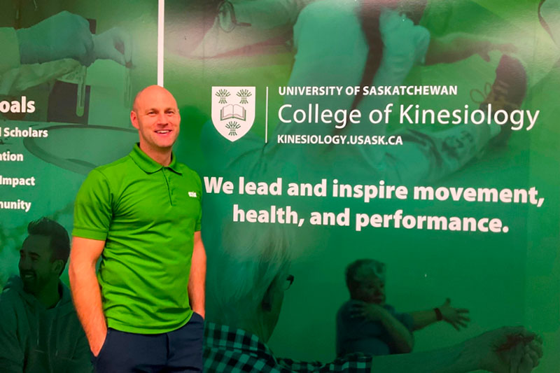 Blair Healey stands in the College of Kinesiology at the University of Saskatchewan in front of a green wall with photos of people stretching