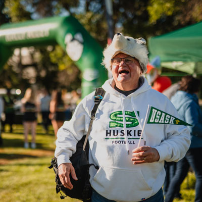 USask grad in white Huskies sweatshirt with a Huskie dog hat and USask pennant